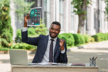 happy african american businessman holding infographics near laptop on deskの写真素材