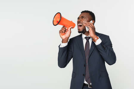 african american businessman in suit screaming in megaphone and talking on smartphone isolated on grayの写真素材