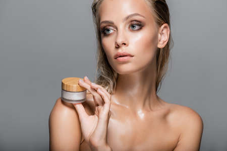 blonde woman with wet skin holding cosmetic cream near bare shoulder isolated on grayの写真素材