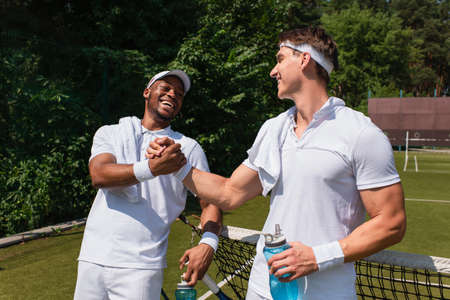 Smiling multiethnic tennis players with sports bottles shaking hands on courtの写真素材