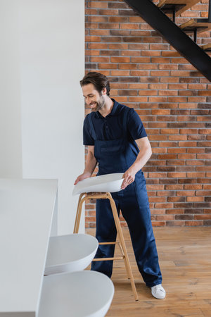 smiling man holding stool near white table in kitchenの写真素材