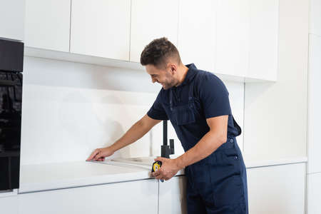 man in overalls measuring tabletop of white furniture in kitchen