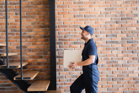 side view of happy mover in uniform walking upstairs with cardboard box