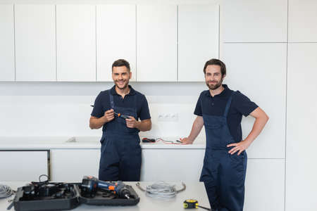 smiling handymen looking at camera near tools on kitchen tableの写真素材