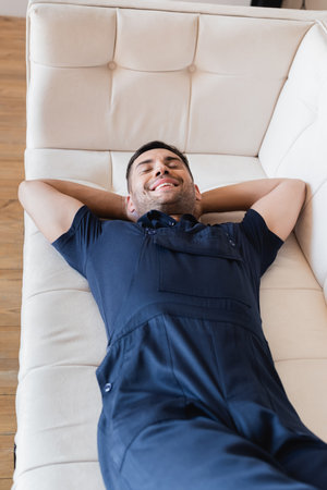 happy worker in overalls relaxing on white couch with closed eyes