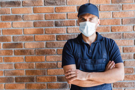 handyman in medical mask standing with crossed arms near brick wall