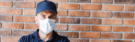workman in cap and medical mask looking at camera near brick wall, bannerの写真素材