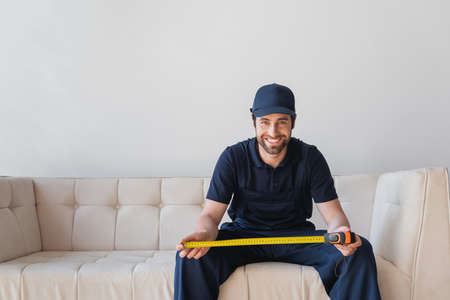 happy worker looking at camera while sitting on white sofa with measuring tapeの写真素材