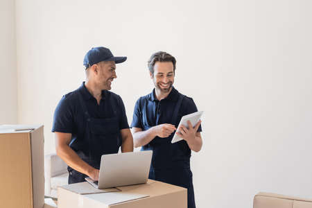 cheerful workman pointing at digital tablet while his colleague typing on laptop near carton boxes