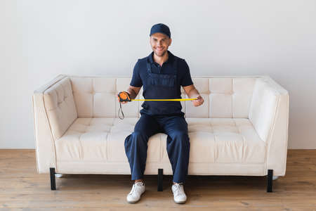 full length view of smiling handyman sitting on white couch with measuring tape