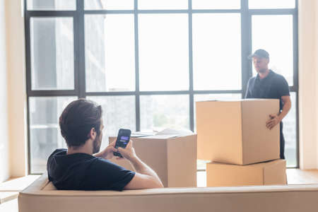 KYIV, UKRAINE - JUNE 24, 2021: mover sitting on couch and messaging on smartphone near blurred...