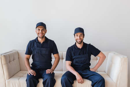 joyful workmen in overalls and caps smiling at camera while sitting on white couchの写真素材