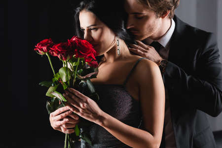 young woman smelling bouquet of red roses near man in suit on black