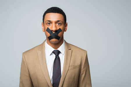 african american businessman with scotch tape on mouth isolated on grayの写真素材