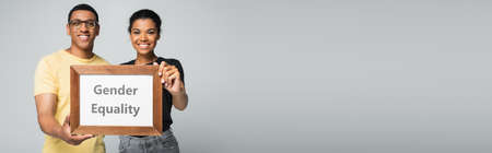 blurred and happy african american man and woman holding wooden frame with gender equality placard isolated on gray, bannerの写真素材