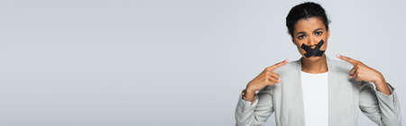african american businesswoman pointing at scotch tape on mouth isolated on gray, banner
