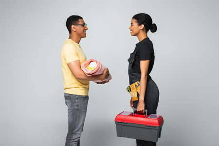 side view of african american father holding baby covered in blanket near repairwoman with toolbox on grayの写真素材