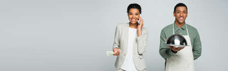 happy african american businesswoman holding dollar banknotes and talking on smartphone near waiter isolated on gray, bannerの写真素材