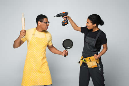 angry african american man in apron holding rolling pin and frying pan while fighting with repairwoman isolated on grayの写真素材