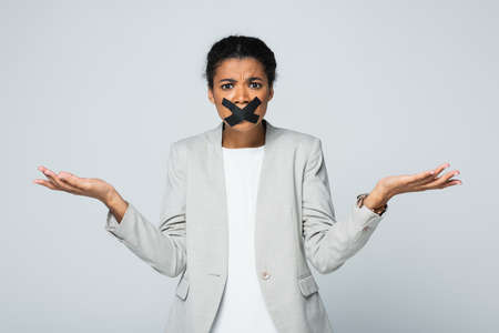 confused african american businesswoman with scotch tape on mouth showing shrug gesture isolated on grayの写真素材