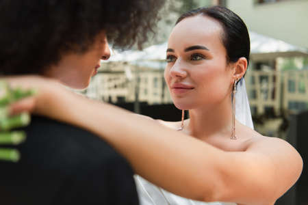 Lesbian woman in wedding dress hugging blurred african american girlfriend outdoorsの写真素材