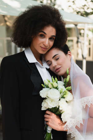 Young lesbian woman in veil holding bouquet near african american girlfriend in suitの写真素材