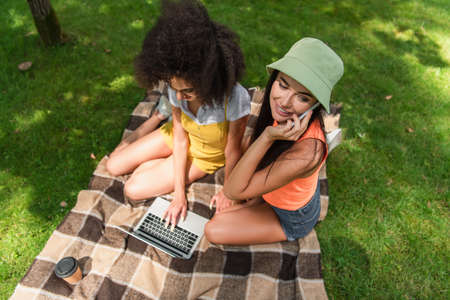 Overhead view of smiling woman talking on smartphone near african american girlfriend using laptop...