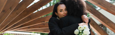 Young lesbian woman holding wedding bouquet and hugging african american girlfriend in park, bannerの写真素材