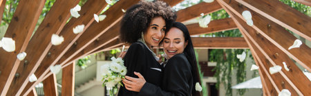 Lesbian african american woman hugging cheerful girlfriend with wedding bouquet under falling petals in park, bannerの写真素材