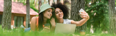 Smiling interracial lesbian couple with coffee taking selfie near laptop in park, banner