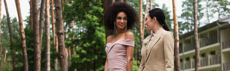 Cheerful interracial lesbian couple in formal wear looking at each other in park, bannerの写真素材