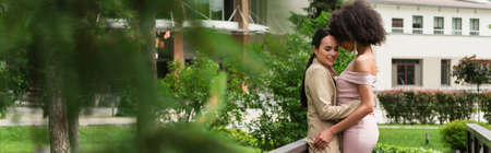 Smiling woman in suit hugging african american girlfriend in dress in park, bannerの写真素材