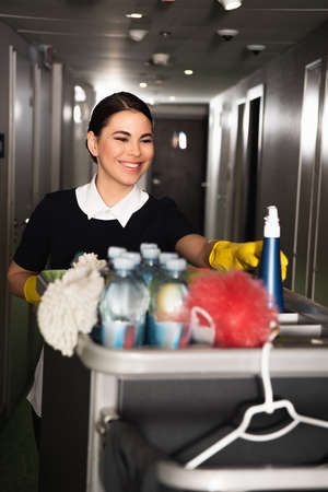 cheerful maid in rubber gloves reaching spray bottle in housekeeping cartの写真素材