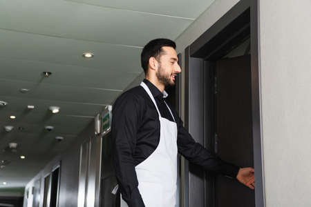 side view of cheerful housekeeper near opened door in hotel roomの写真素材