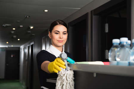young maid in rubber glove reaching mop on housekeeping cartの写真素材
