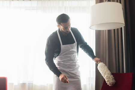 bearded housekeeper cleaning red armchair with dust brushの写真素材