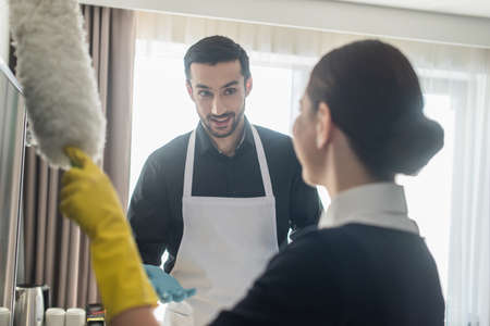 smiling housekeeper in rubber gloves looking at blurred chambermaid with dust brush cleaning tv screenの写真素材