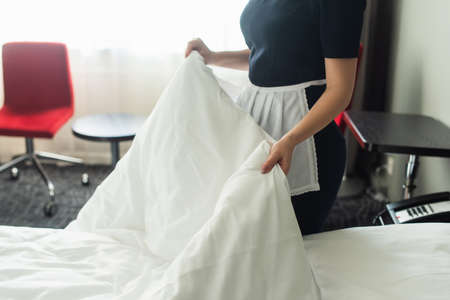 cropped view of young chambermaid in uniform changing bedding in hotel roomの写真素材