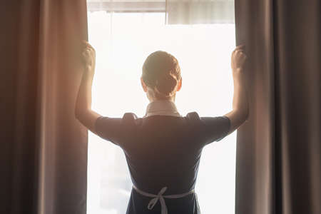 back view of maid adjusting curtains in hotel roomの写真素材