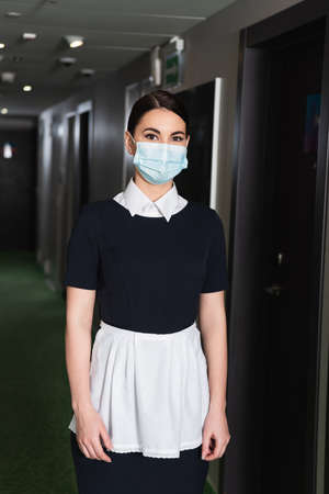 young chambermaid in medical mask and uniform looking at camera in corridor of hotelの写真素材
