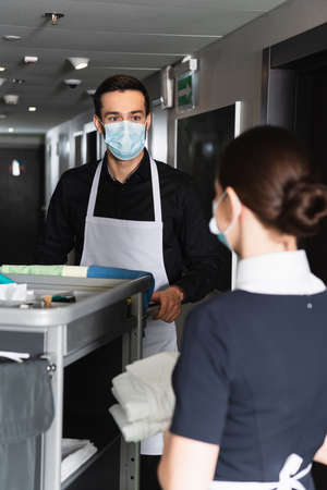 housekeeper in medical mask looking at blurred maid near housekeeping cartの写真素材