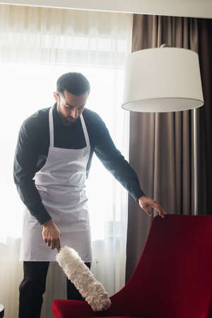 bearded housekeeper in apron cleaning red armchair with dust brushの写真素材