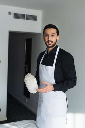 bearded housekeeper in apron holding dust brush on hotel roomの写真素材