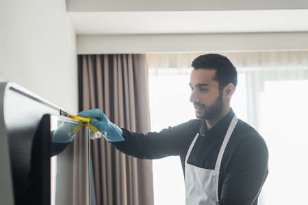 bearded housekeeper in blue rubber glove cleaning tv display with ragの写真素材