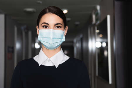 brunette maid in medical mask and uniform looking at camera in corridor of hotelの写真素材