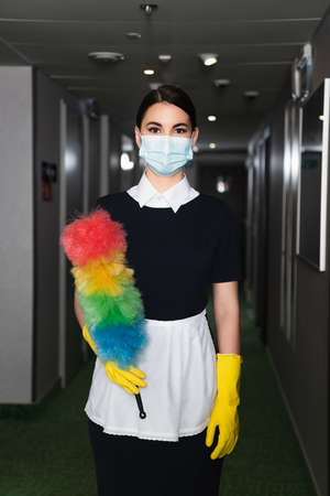 maid in medical mask and rubber gloves holding dust brush in corridor of hotelの写真素材
