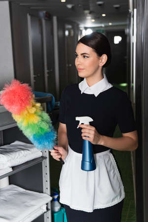 young maid in uniform holding dust brush and spray bottle