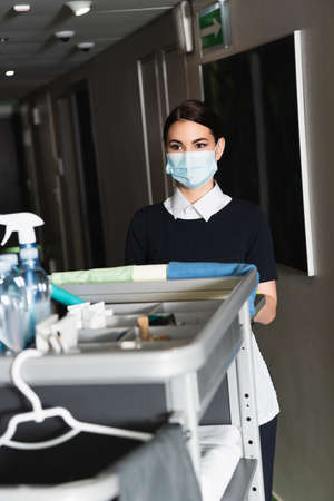 young chambermaid in medical mask standing near blurred housekeeping cartの写真素材