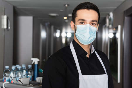 housekeeper in apron and medical mask looking at camera in corridor of hotelの写真素材
