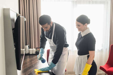 smiling maid looking at colleague cleaning wooden surface with ragの写真素材